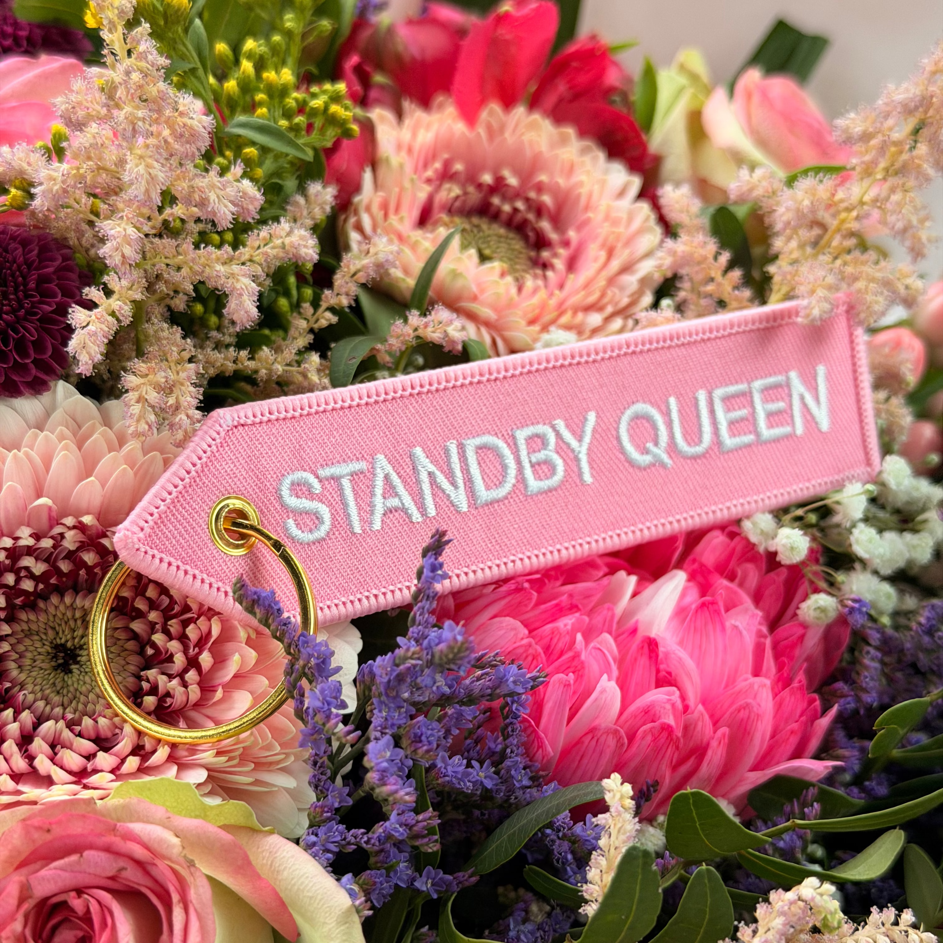 Rosa Schlüsselanhänger Standby Queen vor buntem Blumenstrauss mit goldenem Ring