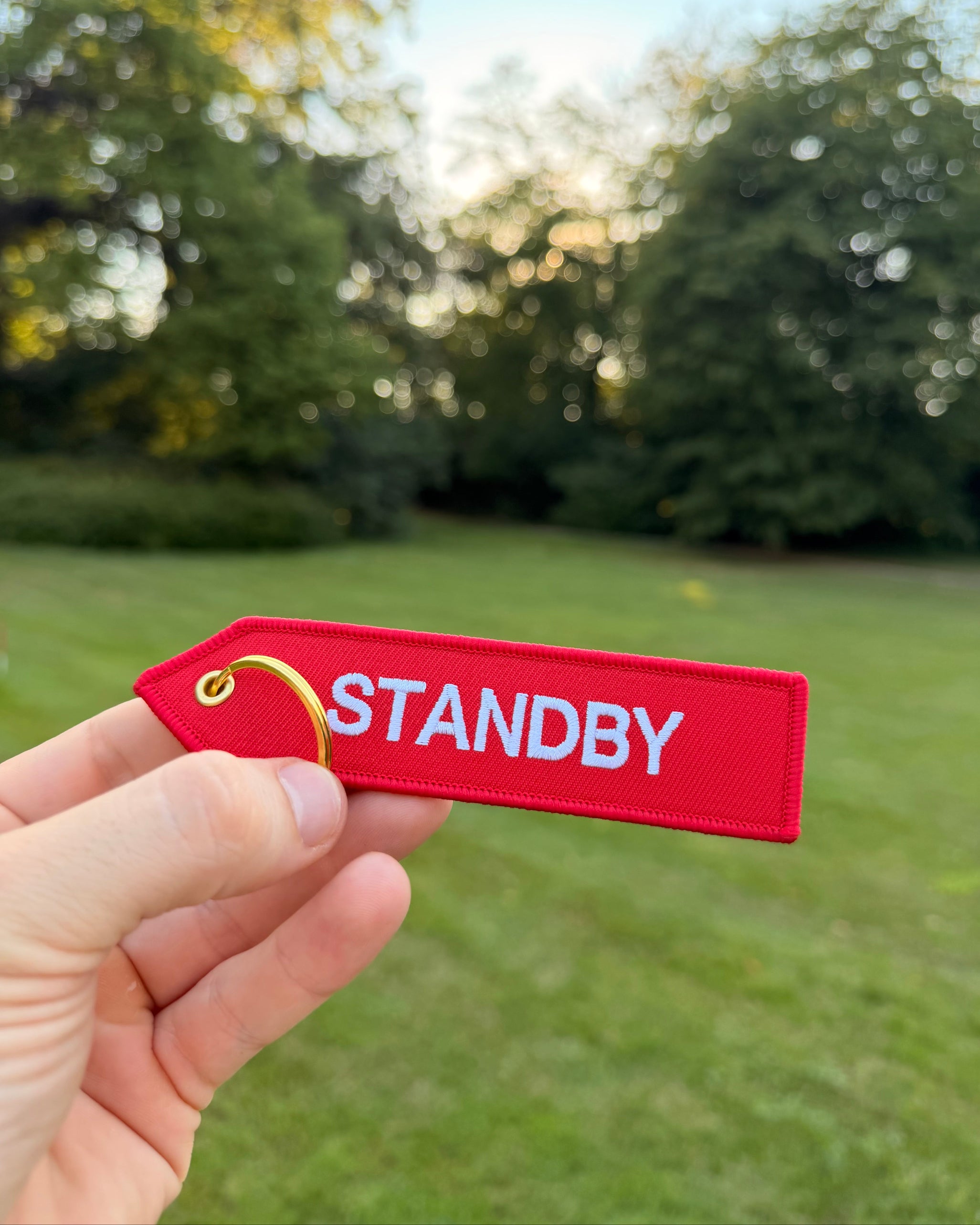 Roter „Standby“ Schlüsselanhänger mit weißer Stickerei und goldfarbenem Ring in einer Hand